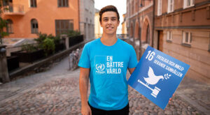 Elias kollar leendes in i kameran där han står på en kullerstensgata och håller i en skyld med globala målen. Han har på sig FN-förbundets ljusblåa t-shirt där det står "En bättre värld".