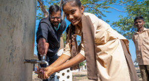 En flicka i beige kläder tvättar händerna i en skola i Indien. I bakgrunden står skolans rektor och vrider på kranen. Solen lyser klart.