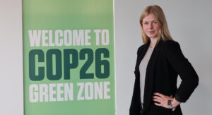 Alicia Nilsson, med svarta kläder och långt blont hår, står vid en grön skylt som det står "Welcome to COP26 Green Zone" på. Hon har ena handen på höften och tittar in i kameran.