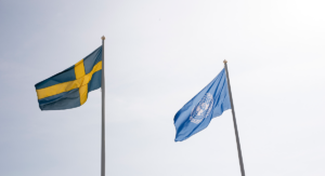 Svenska flaggan och FN flaggan blåser i vinden mot en blå himmel.