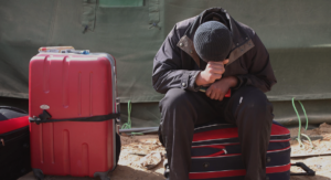 En man sitter på sin resväska framför ett tält, han lutar huvudet bedrövat ner i sina händer