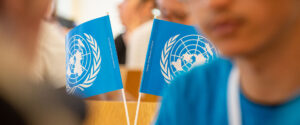 Två små bordsflaggor med FN:s logga syns i bakgrunden. I förgrunden syns några ungdomar i oskärpa som tycks lyssna på någon.